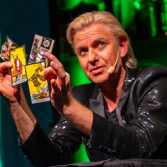 En person utför ett trolleritrick eller en tarotläsning, håller flera kort utspridda i handen på en grönbelyst scen.