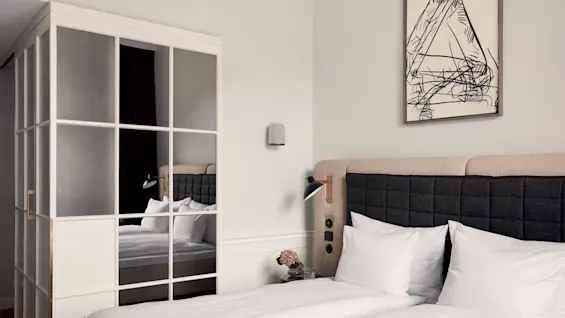 A neatly made bed with white sheets and pillows stands next to a mirrored wardrobe in a modern bedroom. A sketch adorns the wall above the headboard. 