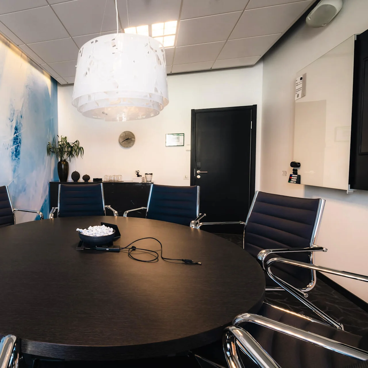 Ett runt konferensbord med stolar står under en stor vit taklampa. Rummet har en blå abstrakt vägg, modern inredning och en svart dörr.