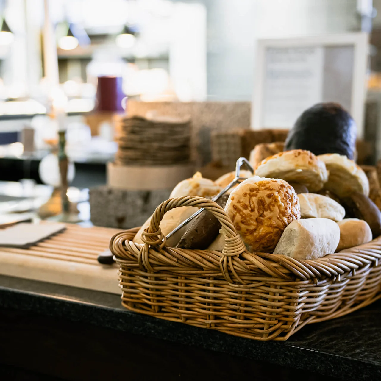 En flettet kurv fyldt med forskellige brød står på en disk. I baggrunden er en buffet med tallerkener og slørede køkkendekorationer.