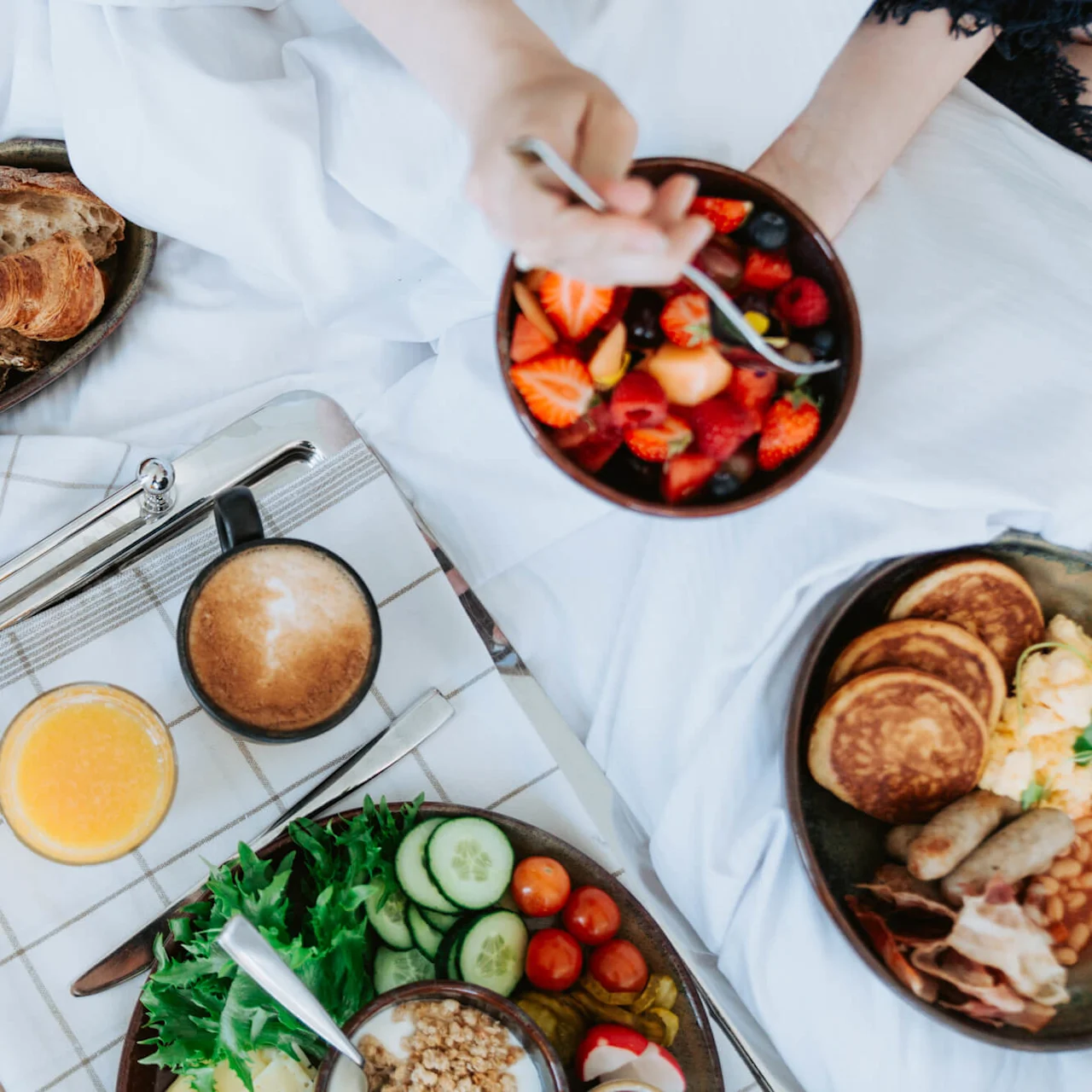 Breakfast in bed features fresh fruit, croissants, and an assortment of pastries and breads on a plaid cloth, accompanied by coffee and orange juice, creating a cozy and inviting setting.
