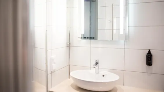 En vit keramisk vask med modern kran är monterad på en badrumsbänk. En spegel ovan visar partiella reflektioner, med texten "IT'S GETTING HOT IN HERE". Vita kaklade väggar ger ett rent sammanhang.