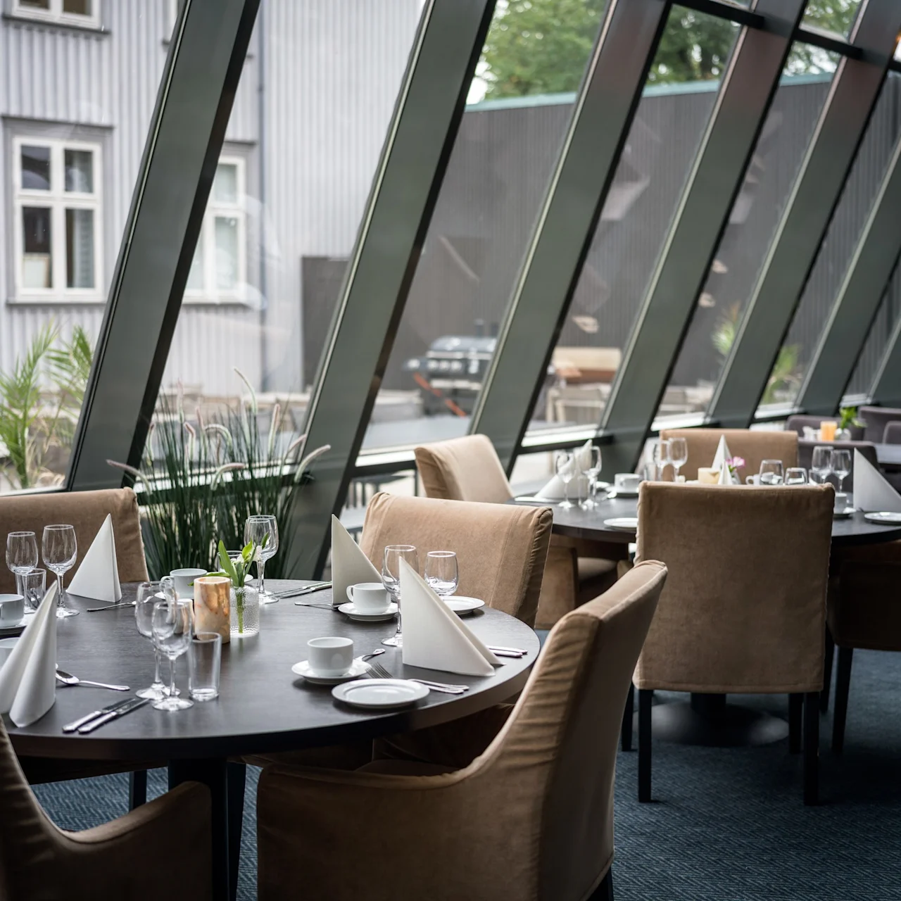 Eleganta bord dukade med vita servetter och glas; bekväma stolar omger dem i en modern, glas-inhägnad restaurang med utsikt över en stadsbild med nära växter.