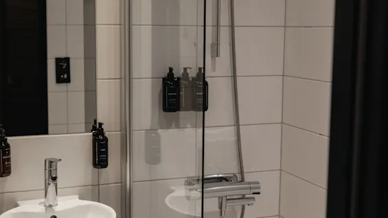 A clean, modern bathroom featuring a sink, mirror, and a bathtub with an overhead shower. Dispensers for body wash, shampoo, and conditioner are visible.