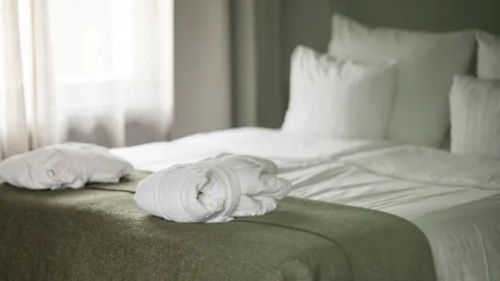 Kutsuva hotellisänky raikkailla valkoisilla liinavaatteilla ja tyynyillä, jota korostavat vihreä viltti ja kaksi pehmeää kylpytakkia, kutsuen rentoutumaan.