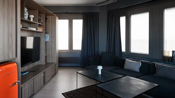 A modern living room with a grey sofa and black square tables under large windows. An orange fridge is next to a shelving unit holding books and decor items. 