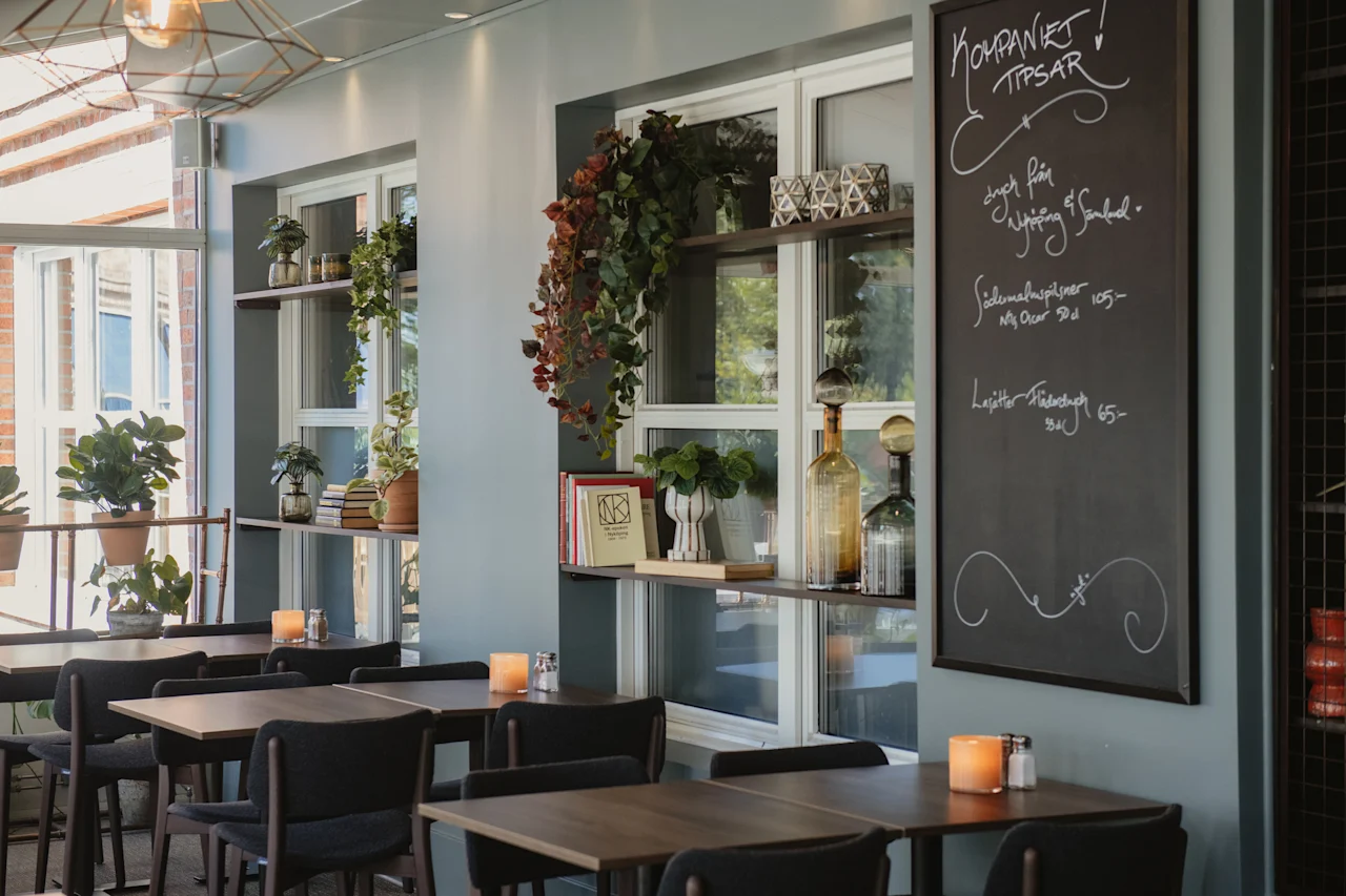 En ljus kaféinteriör med dukade bord, gröna växter och en griffeltavla. Synlig text: KOMPANIET TIPSAR, Södermalmspilsner 105, Lajitas Fläderdryck 65.