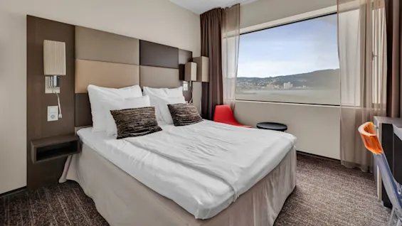 A neatly made bed with white linens and decorative pillows stands in a modern room, facing a large window with a view of a waterfront cityscape. 