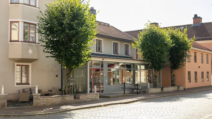 En lys dag på en brosteinsgate, kantet med bygninger og grønne trær. En bygning har en glassfasade med MAJOREN.