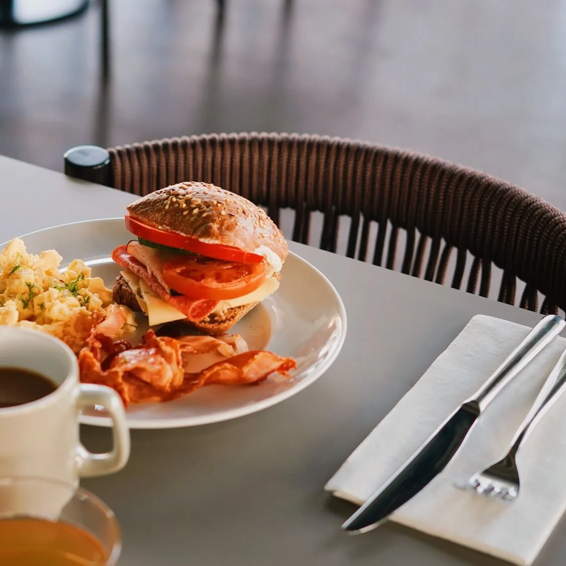 A breakfast plate with a sandwich, scrambled eggs, and bacon sits on a table. Nearby are cups of coffee and orange juice, with a fork and knife on a napkin.