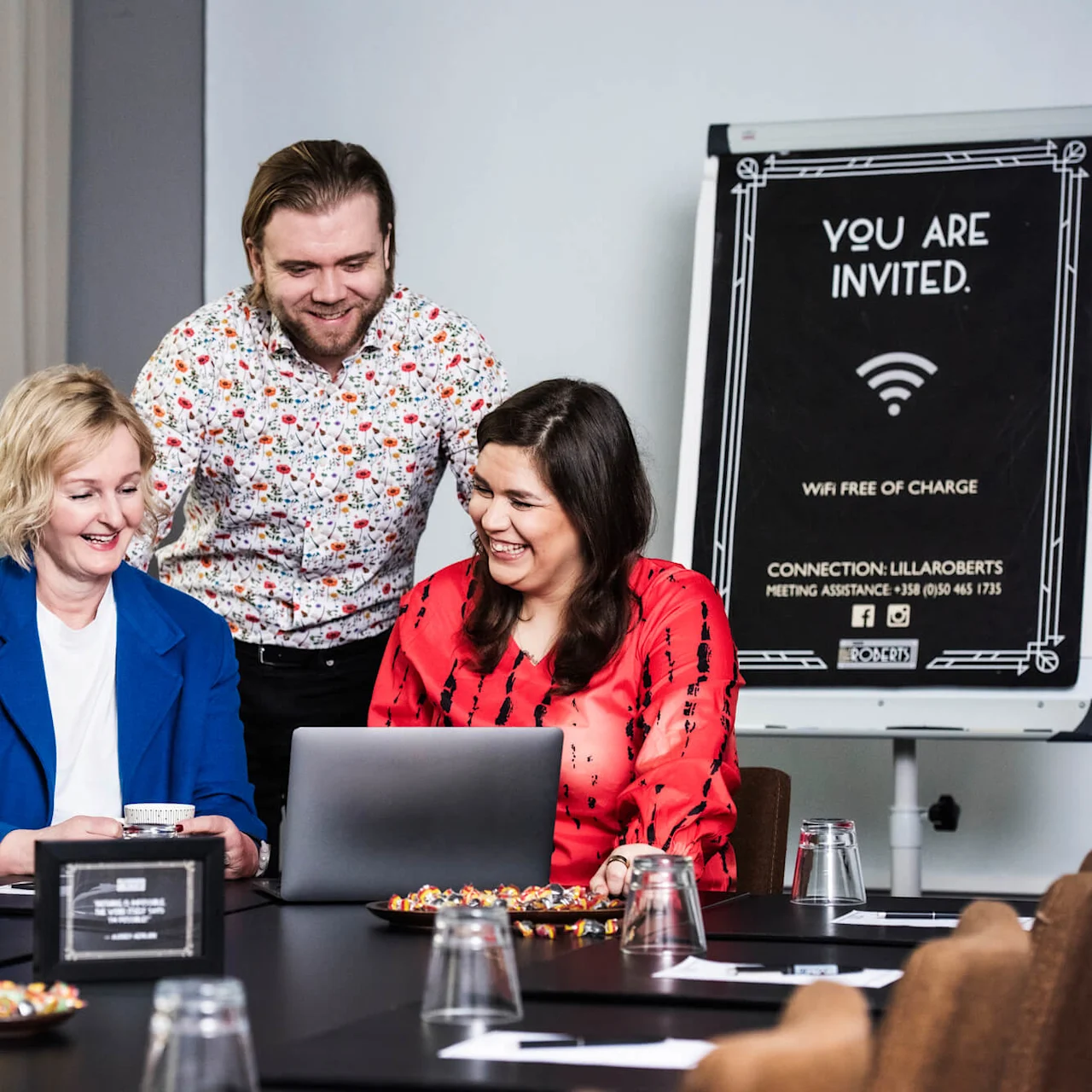 Tre personer smiler mens de er sammen om en laptop på et møtebord, omgitt av notatbøker og snacks. Et skilt sier: "YOU ARE INVITED. WIFI FREE OF CHARGE. CONNECTION: LILLAROBERTS MEETING ASSISTANCE +358 (0)50 465 1735."