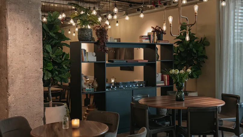 Café with wooden tables and chairs, softly lit by hanging bulbs. Shelves with books and plants divide seating areas. Greenery decorates the cozy, dimly-lit space with a relaxed atmosphere.