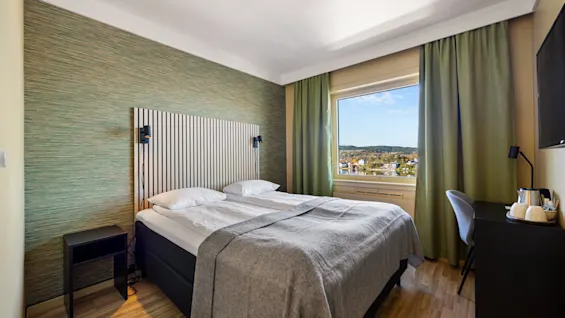 Lyst hotellrom med dobbeltseng, grønn fondvegg og vindu med utsikt.