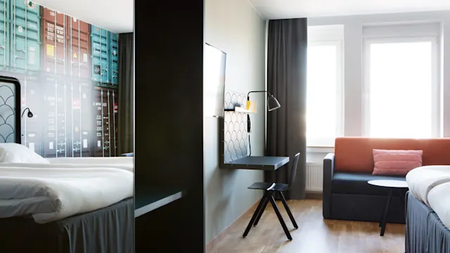 Dubbelsängar är prydligt arrangerade i ett modernt hotellrum. Ett skrivbord med en väggmonterad TV vetter mot sängarna, kompletteras av en soffa och fönster som släpper in naturligt ljus.