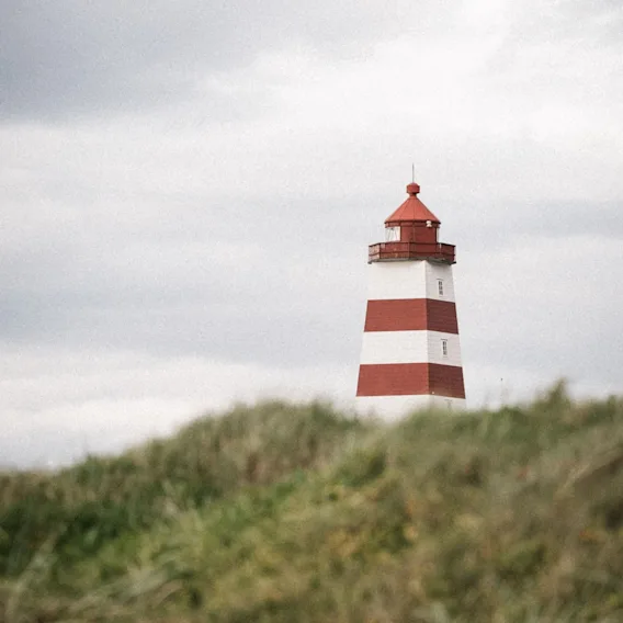 Det rød- og hvitstripete Alnes fyr bak gresskledde sanddyner under en skyet himmel.