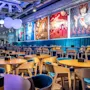 Stylish restaurant interior features circular wooden tables and turquoise chairs, beneath large round lights. Colorful pop art posters adorn the walls, creating a contemporary, vibrant atmosphere.