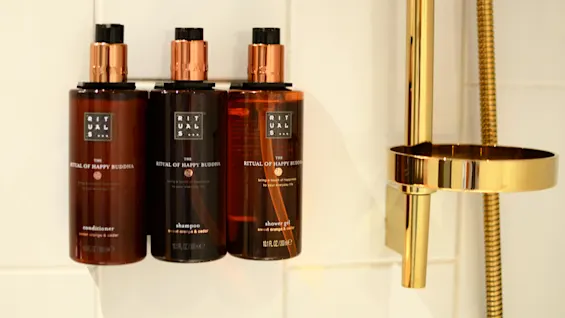 Three brown bottles labeled "The Ritual of Happy Buddha" with a conditioner, shampoo, and shower gel, on a white tiled shelf.