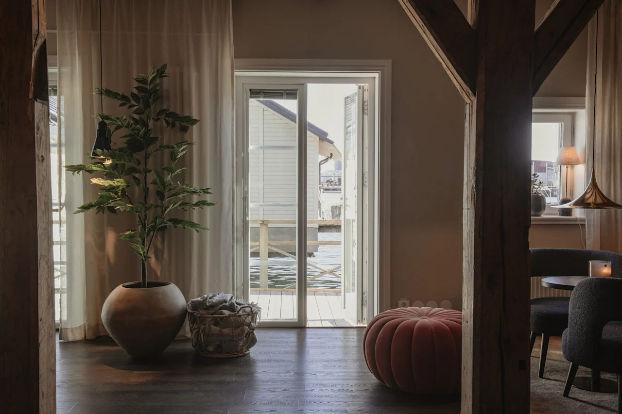 Et hyggeligt værelse med en stor potteplante og en rund puf, med udsigt gennem en åben dør til en vandudsigt med en bygning.