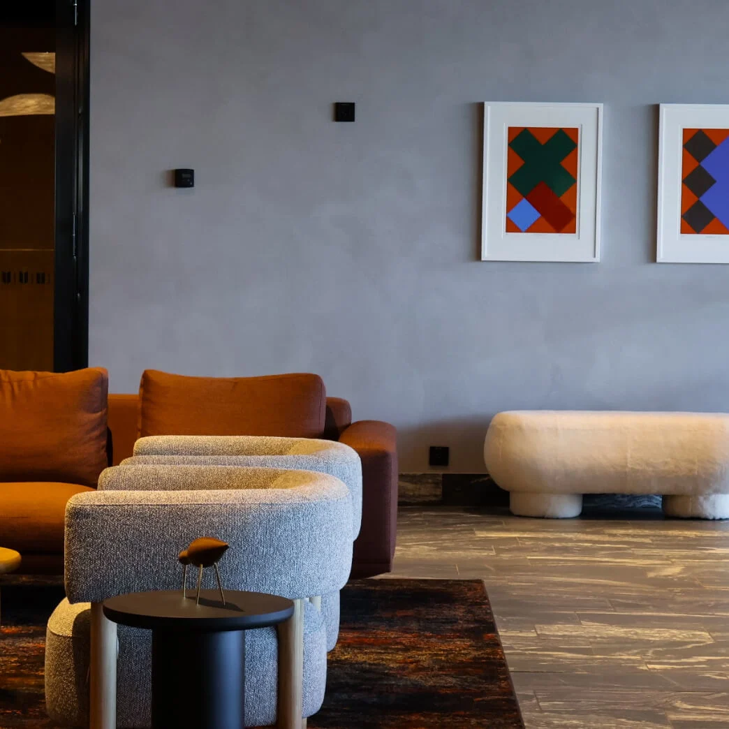 Brown sofa sits in a modern lounge with gray chairs and round tables; abstract art hangs on a gray wall alongside a standing lamp, creating a cozy atmosphere.