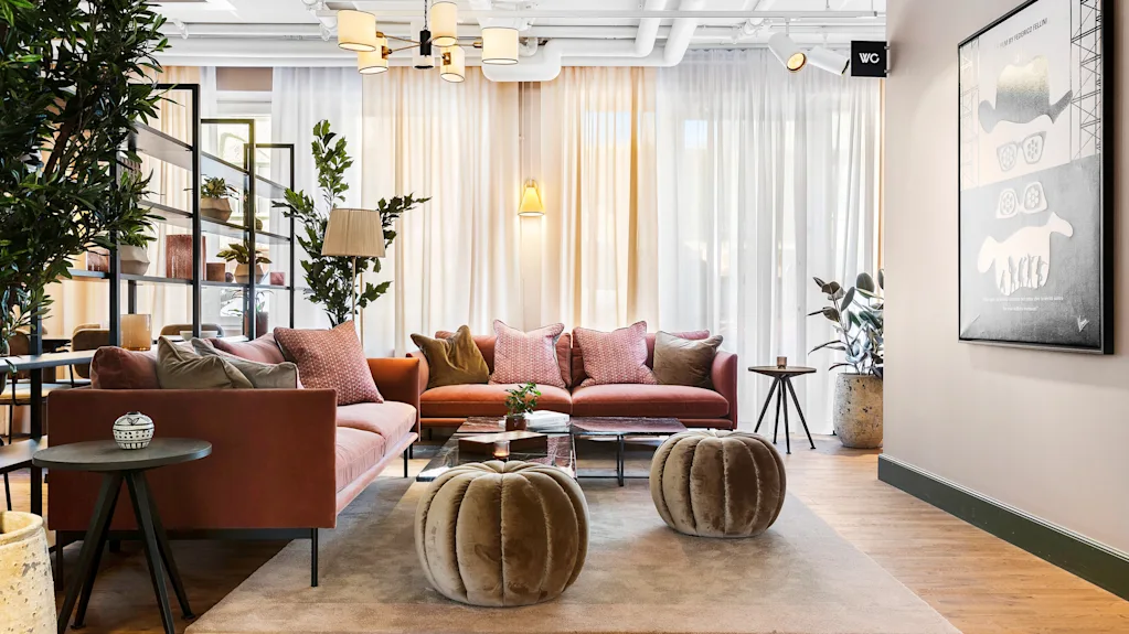 Moderne og koselig hotellobby med rosa sofaer, myke puffer og grønne planter under lunt lys.