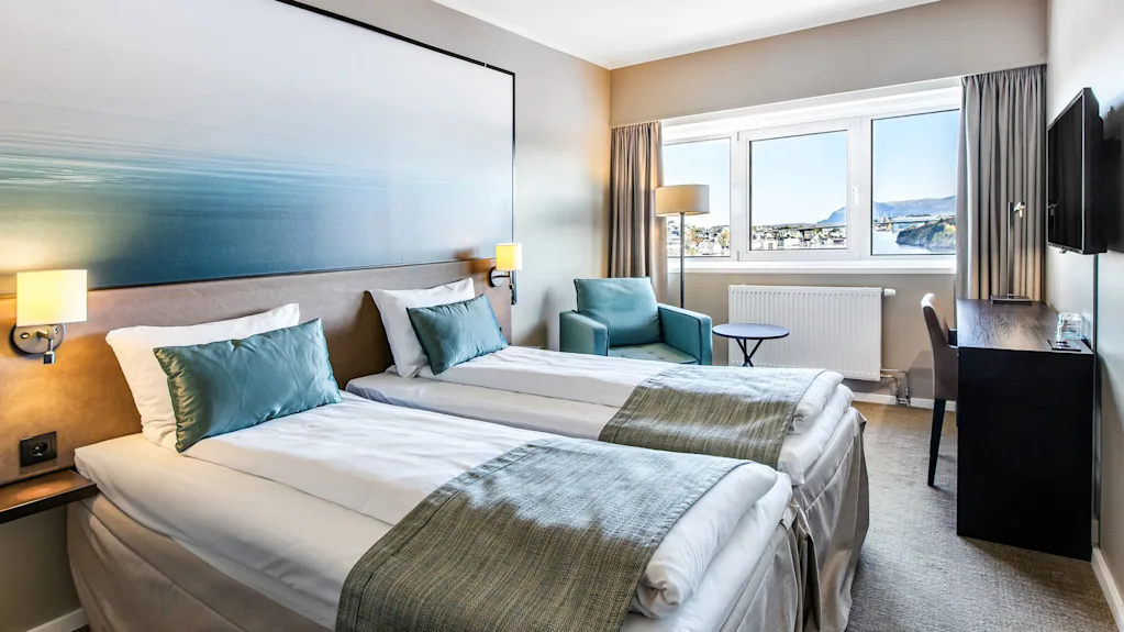 Two neatly made beds with teal pillows and throws, positioned in a bright hotel room. There’s an armchair, small table, and a desk facing large windows offering a scenic city view.