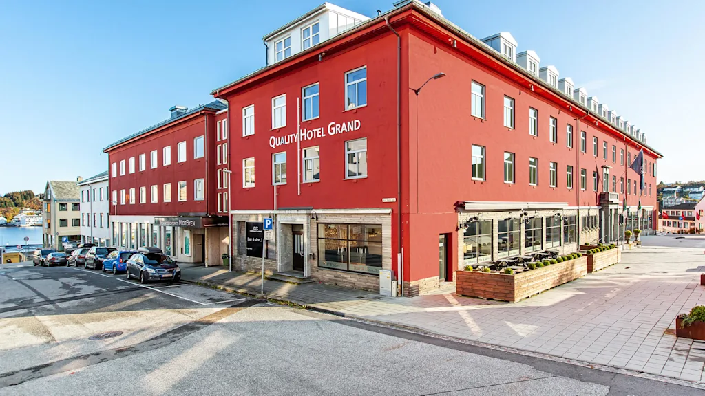 Quality Hotel Grand, en stor rød bygning, står på en gate med parkerte biler og klar himmel. Nær vann indikerer en kystnær setting.