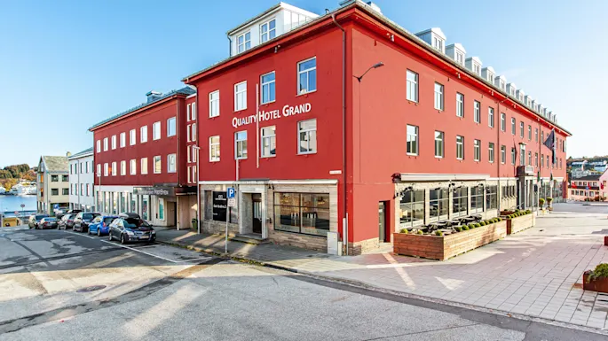 Quality Hotel Grand, a large red building, stands on a street with parked cars and clear skies. Nearby water indicates a coastal setting.