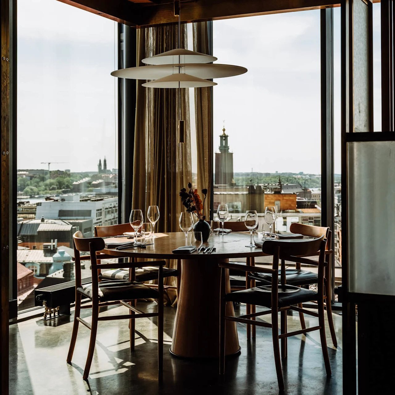 A round wooden dining table is set with wine glasses and cutlery, overlooking a cityscape through large windows. Modern pendant light hangs above, next to a leather bench.