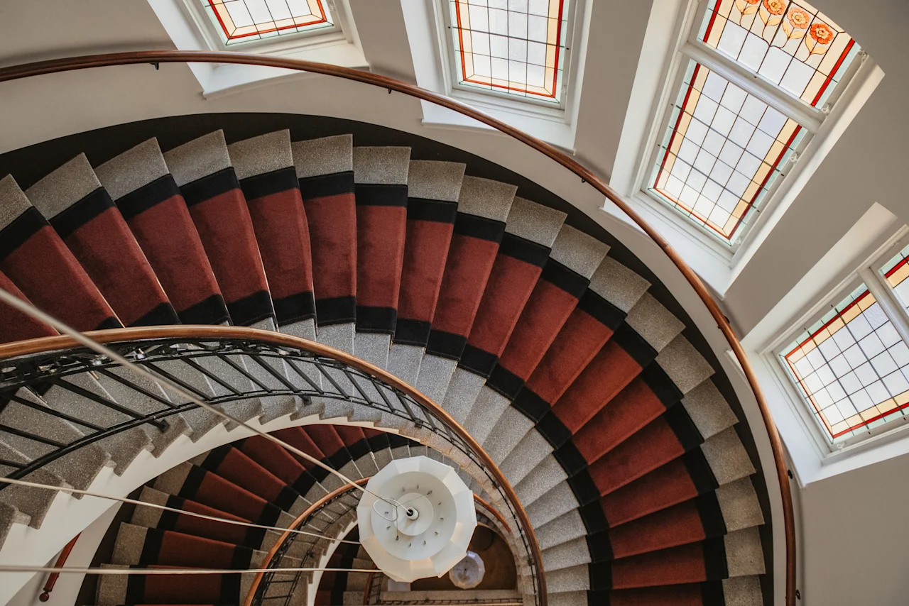 En storslagen spiraltrappa med röd matta och utsmyckade blyinfattade fönster, som leder uppåt i en elegant byggnad.