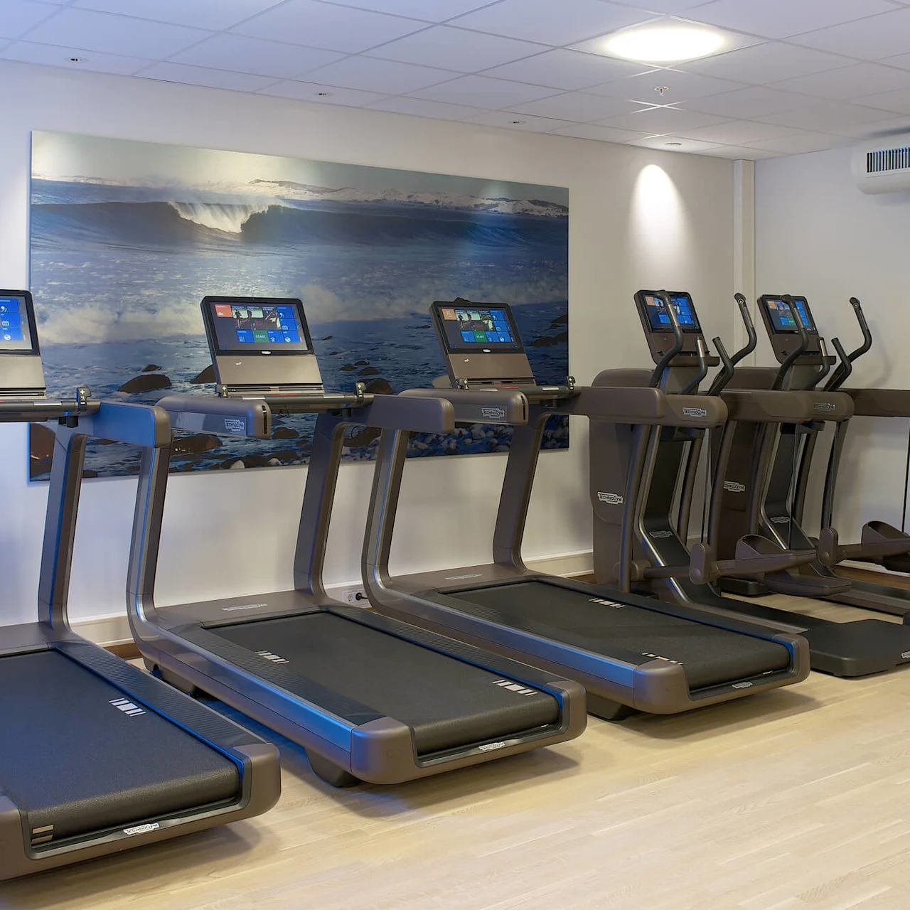 Löpband och elliptiska maskiner står uppradade mot en gymvägg med ocean-tema målningar. Varje maskin har en integrerad digital skärm. Gymmet har en ljus, rymlig atmosfär.