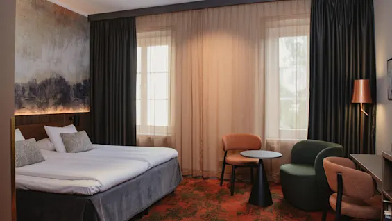 Et innbydende hotellrom med en komfortabel seng, kunstnerisk veggdekor og et koselig hjørne med stoler og et bord.