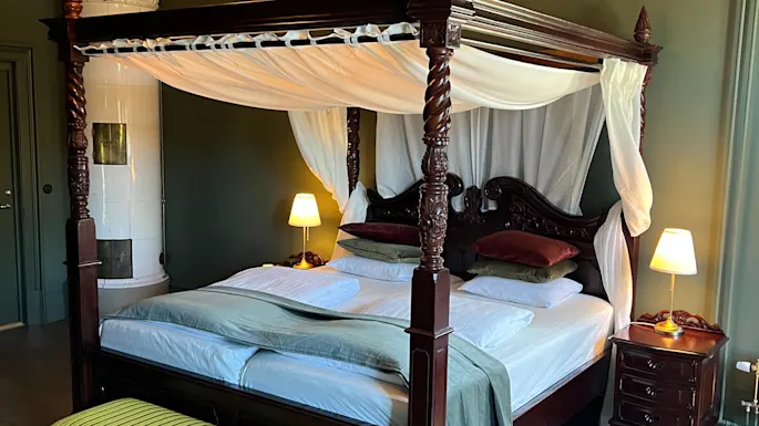 En himmelseng med hvite draperier og grønt sengetøy står sentralt i et elegant rom. En lysekrone henger over, omgitt av grønne vegger og tregulv. Ved siden står en tent lampe og en utsmykket benk.