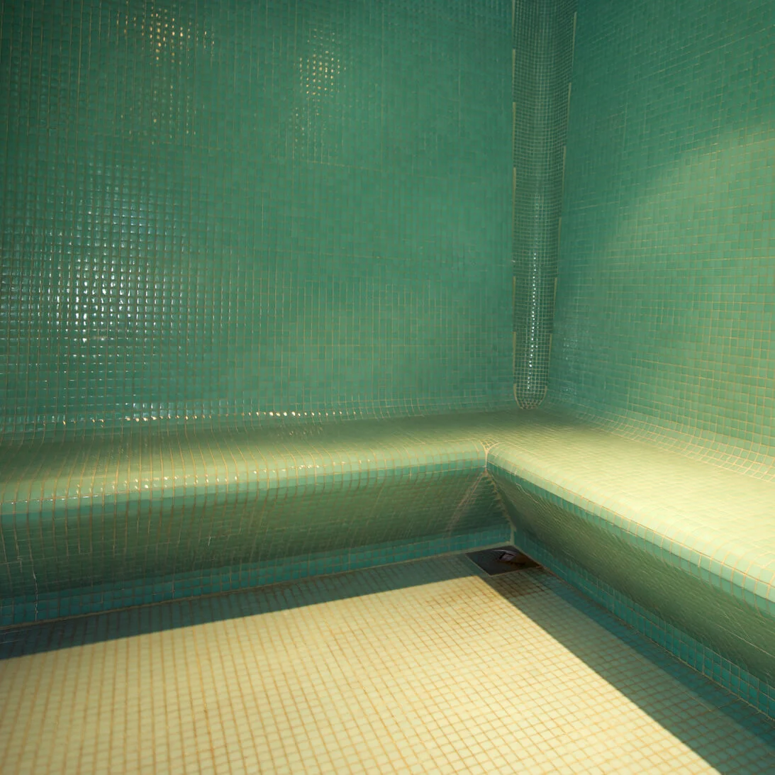 An empty steam room lined with green tiles, with built-in benches and a floor drain.