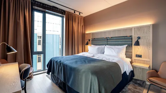 En dobbeltseng med hvidt sengetøj og gråt tæppe er i et moderne hotelværelse, med en træhovedgærde, skrivebord og stort vindue med brune gardiner.