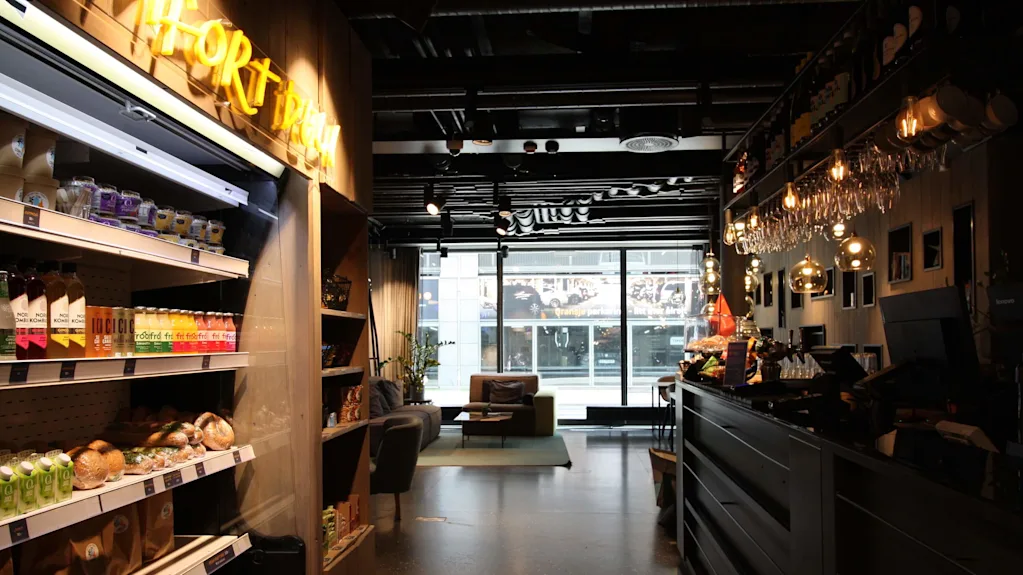 Hylla med drikkevarer, snacks og bakverk ved bar med hengende lamper og kasse; en koselig sofa er synlig gjennom store vinduer. Tekst: "GRAB & GO."