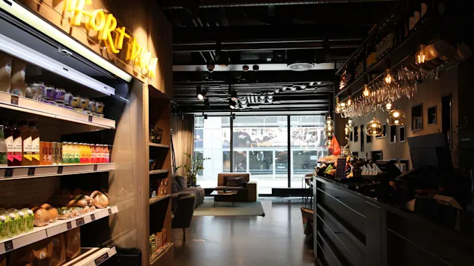 Shelves display drinks, snacks, and baked goods beside a bar with hanging lights and a cash register; a cozy seating area with sofas is visible through large windows. Text: "GRAB & GO." 