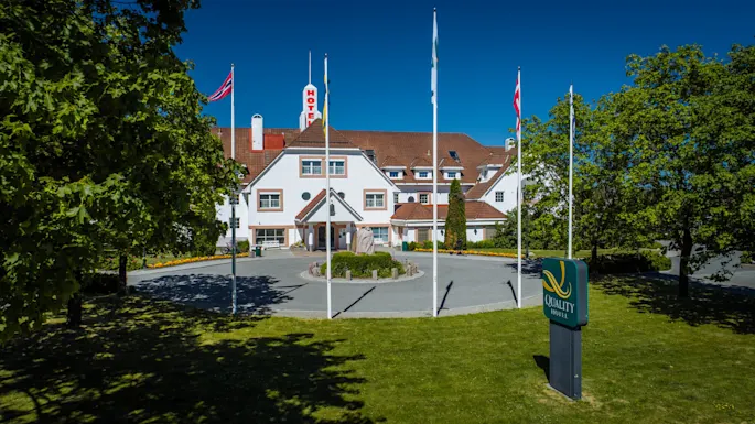 A white multi-story hotel with red "HOTELL" sign, surrounded by trees and flags, stands in sunny weather. A circular driveway leads to the entrance. "QUALITY HOTEL" is on a green sign.