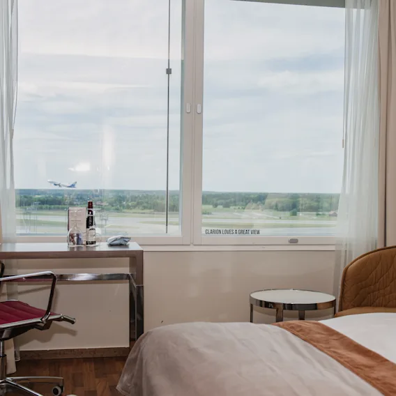 Et hotellrom med skrivebord, stol og seng, som tilbyr en dynamisk utsikt over et fly som tar av fra en rullebane. Vindutekst: LOVES A GREAT VIEW.