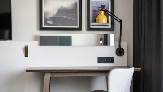 A simple wooden desk holds a gray chair, flanked by a modern wall lamp. Two framed pictures adorn the wall, with a black curtain to the right.