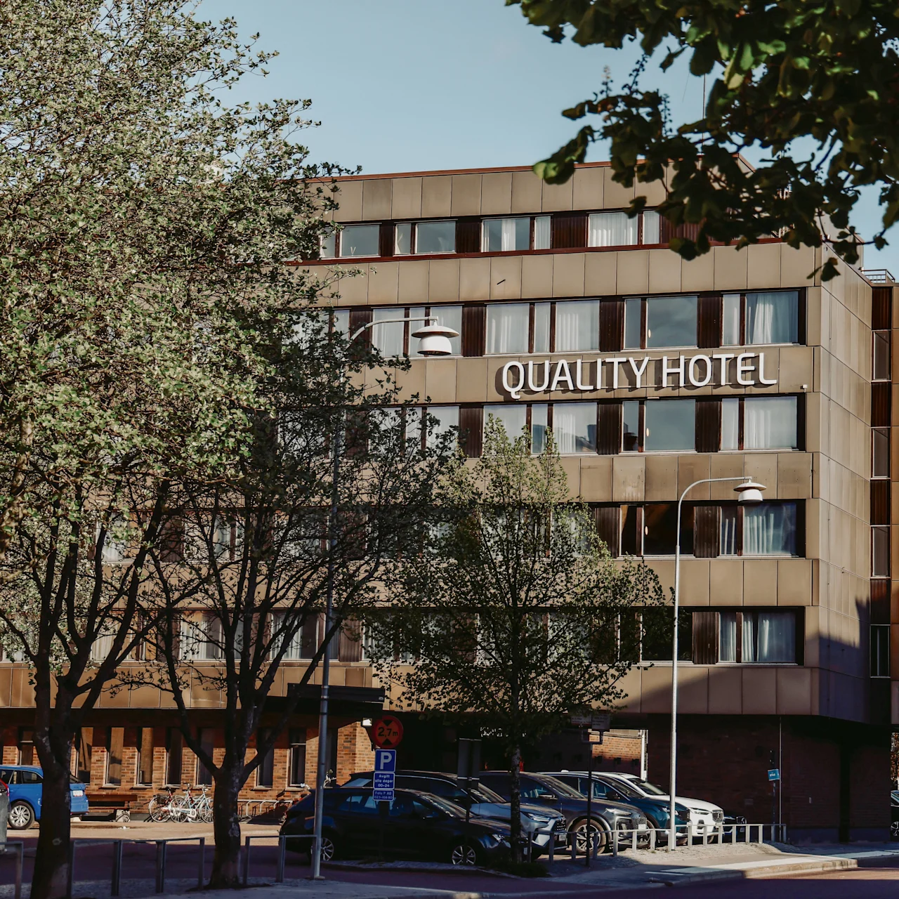 Quality Hotel sign on a modern, multi-story building surrounded by trees and parked cars. The trees are partly obscuring the view of the hotel, creating a natural urban setting.