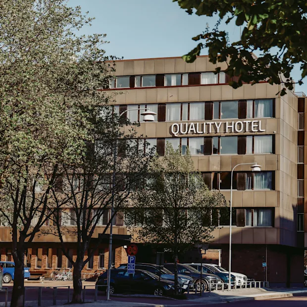Quality Hotel sign on a modern, multi-story building surrounded by trees and parked cars. The trees are partly obscuring the view of the hotel, creating a natural urban setting.