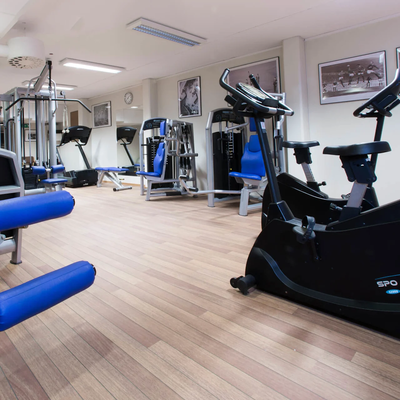 Träningsutrustning i ett gym, med vikter och konditionsmaskiner, på ett trägolv.