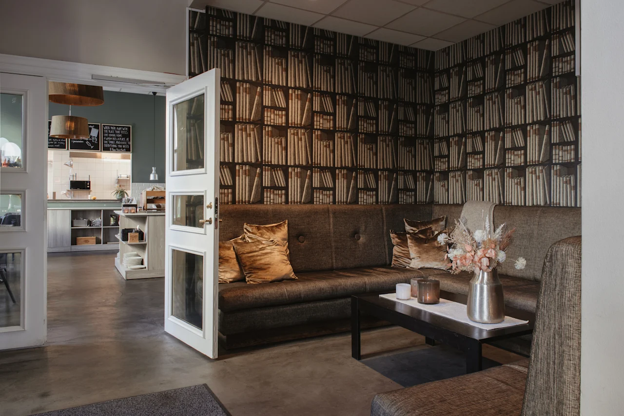 Mysig loungeavdelning med soffa, soffbord och tapet med bokhyllsmotiv, med utsikt mot ett kök. Home Hotel Post.