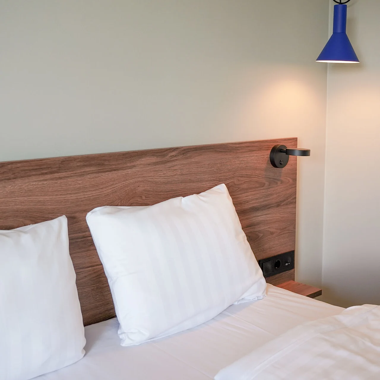 A neatly made bed with white pillows rests against a wooden headboard. A blue wall lamp is mounted above, softly illuminating the minimalist bedroom.