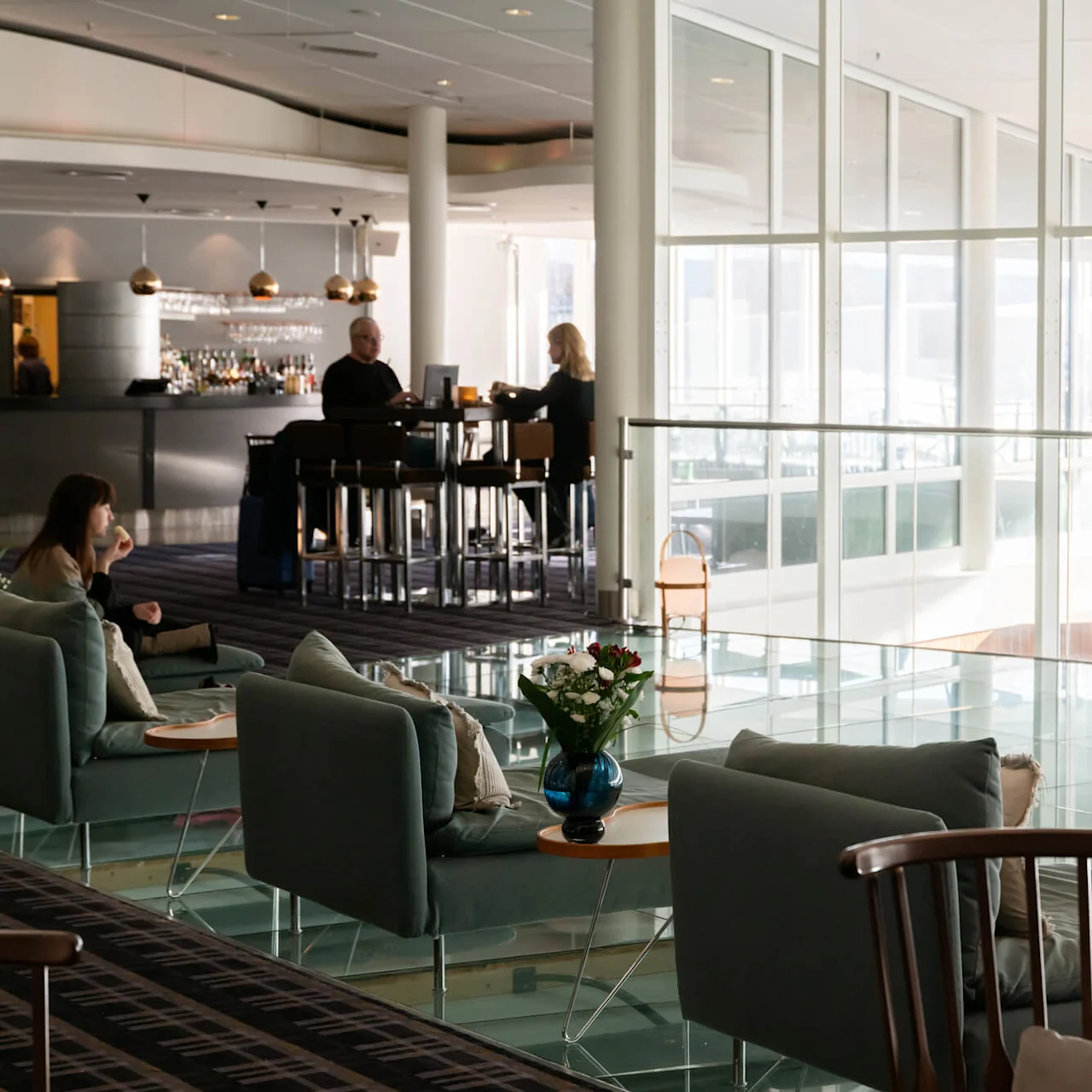 Komfortable loungestoler med puter er plassert nær glassvegger, mens en kvinne drikker kaffe. Bak er en moderne bar med sittende personer synlig i et lyst miljø.