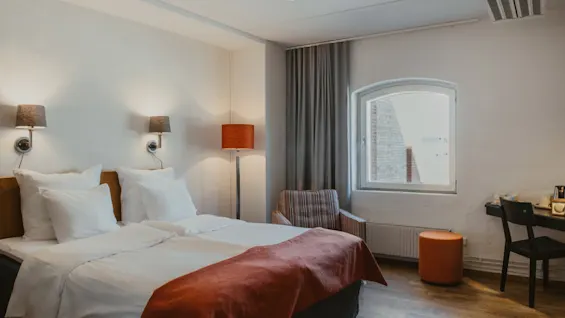 Viihtyisä hotellihuone, jossa on mukava sänky, ikkuna, raidallinen nojatuoli ja pieni työpöytä tuolineen.