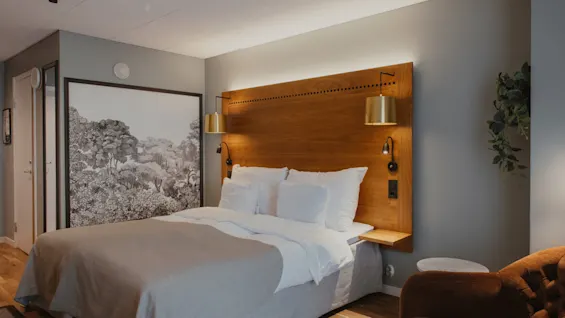 Et hyggeligt hotelværelse med en stor seng med træhovedgærde, stilfulde lamper og et naturinspireret kunstværk. En lænestol er synlig.