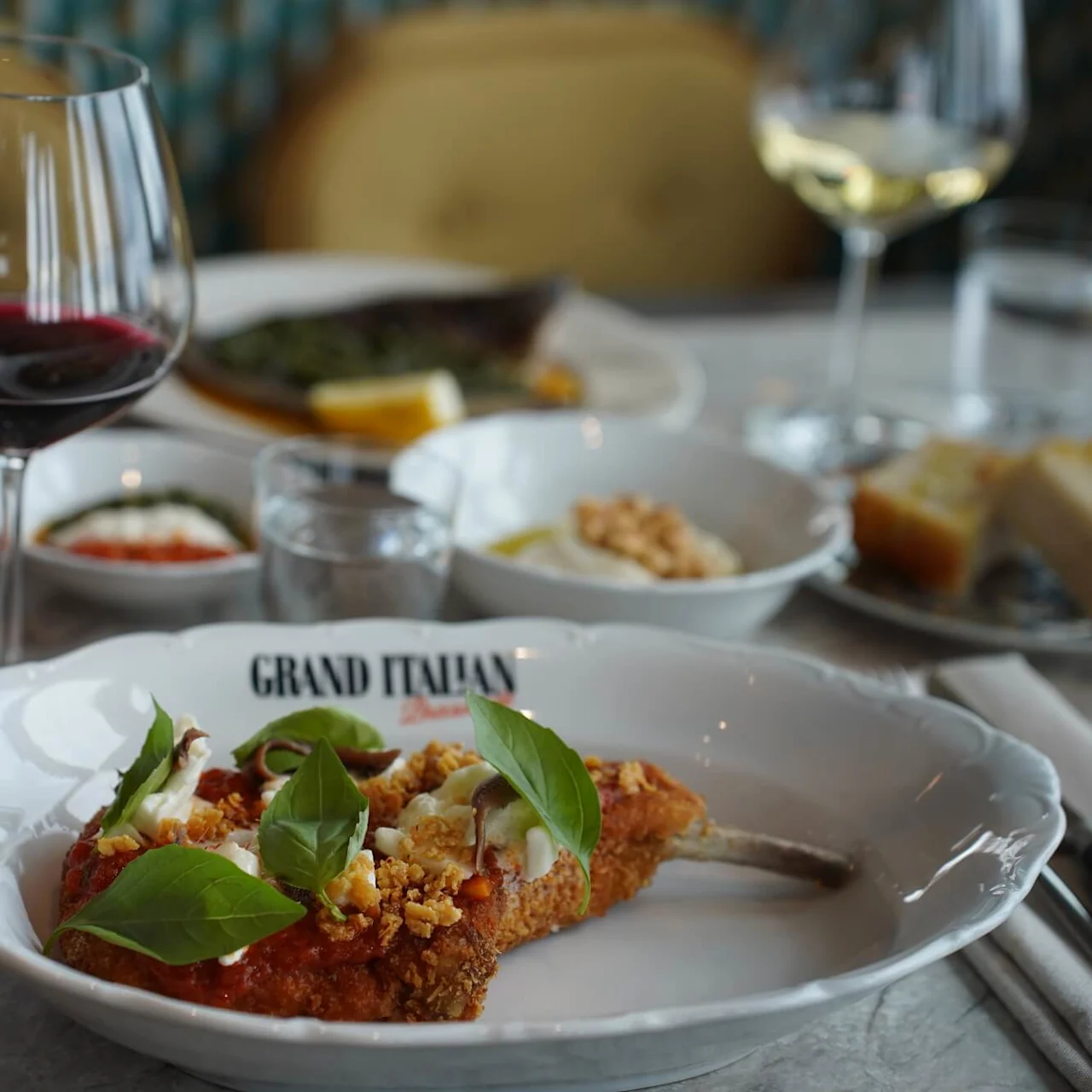 En tallerken med panert kjøtt toppet med basilikum og saus står på et bord. Teksten på tallerkenen lyder "GRAND ITALIAN." Vinglass og forskjellige retter omgir den.
