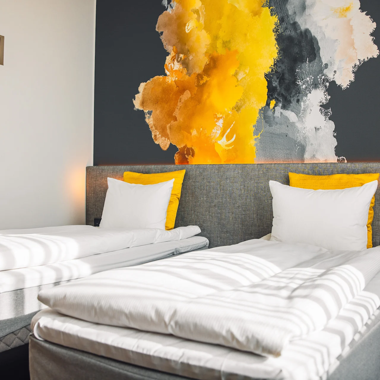 Two neatly made single beds with white linens and yellow accents, positioned against a wall featuring a vibrant abstract mural, in a brightly lit modern room. 
