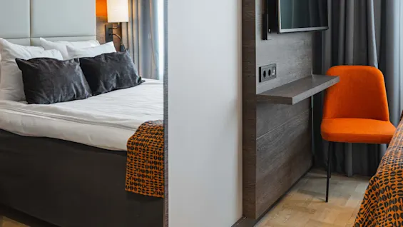 En modern hotellrum med en säng till vänster och en väggmonterad TV till höger. En orange stol är under TV:n bredvid grå gardiner, kompletterad av ett trägolv.